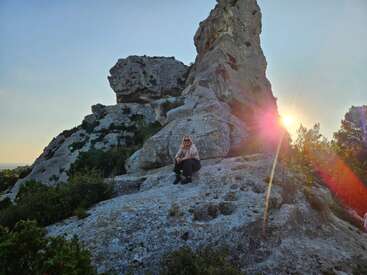 A person sits on a large rock formation at sunset, surrounded by rugged cliffs and greenery. Sunlight creates a lens flare, adding warmth and vibrancy.