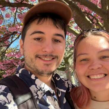 A young man and woman smile for a selfie under blooming pink cherry blossom trees on a sunny day, enjoying nature and each other's company outdoors.