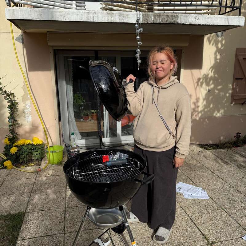 A person stands on a patio, smiling with closed eyes, holding a black barbecue grill lid. Sunlight shines, plants and household items surround them. Casual, relaxed setting.