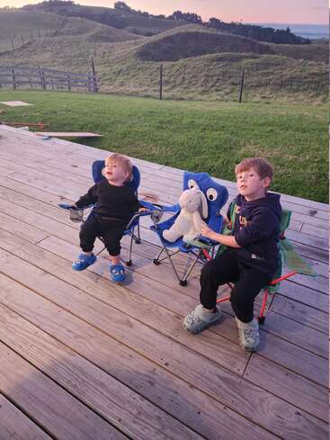 Zwei kleine Jungen sitzen auf blauen Klappstühlen auf einer Holzterrasse und blicken nach oben. Einer hält ein Stofftier in der Hand. Im Hintergrund sind grüne Hügel und ein Sonnenuntergang zu sehen.