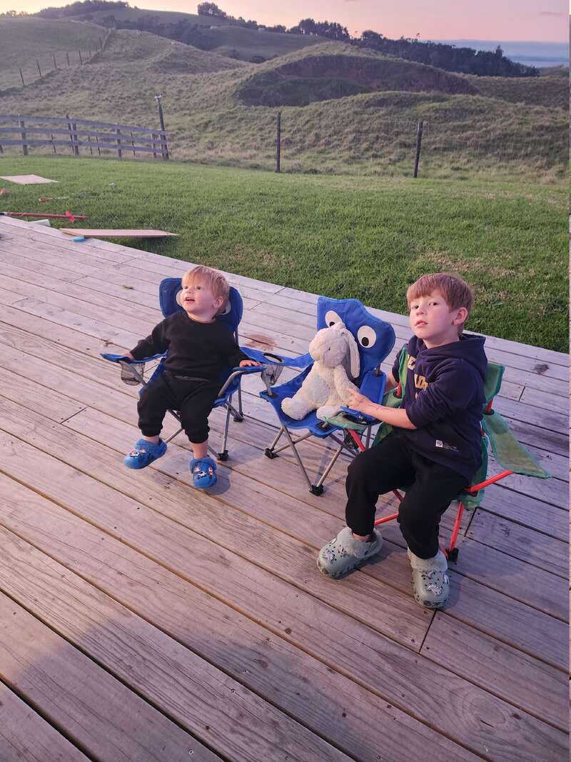 Zwei kleine Jungen sitzen auf blauen Klappstühlen auf einer Holzterrasse und blicken nach oben. Einer hält ein Stofftier in der Hand. Im Hintergrund sind grüne Hügel und ein Sonnenuntergang zu sehen.
