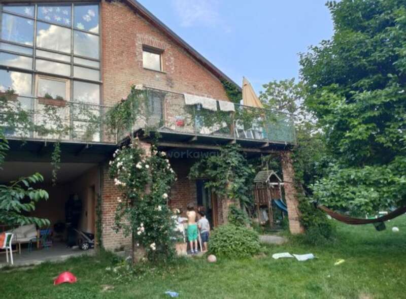 L'image représente une maison en briques à deux étages avec un balcon, entourée d'une végétation luxuriante et d'un jardin animé, avec des enfants jouant dans la cour.