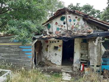 Das Bild zeigt eine baufällige Holzkonstruktion mit abblätternder Farbe und einer fehlenden Tür, die auf einer Wiese mit Bäumen im Hintergrund steht.