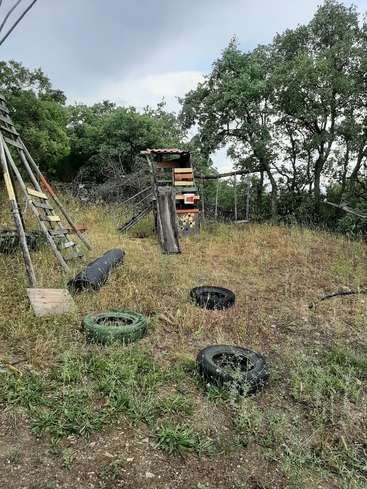 Das Bild zeigt eine Grasfläche mit einer Holzkonstruktion, einer Leiter und verstreuten Reifen, umgeben von Bäumen unter einem bedeckten Himmel.