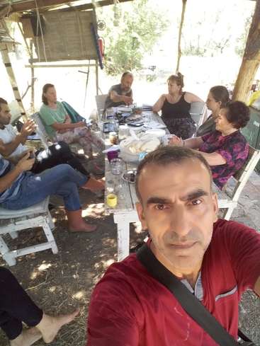 Das Bild zeigt eine Gruppe von Menschen, die um einen Tisch im Freien sitzen, während eine Person im Vordergrund ein Selfie macht.