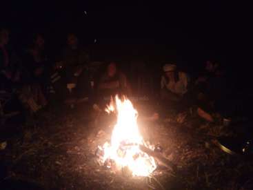 Das Bild zeigt eine Gruppe von Menschen, die sich nachts um ein Lagerfeuer versammeln, in dessen Mitte das Feuer hell brennt.