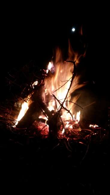 Das Bild zeigt ein lebhaftes Lagerfeuer mit hoch aufsteigenden Flammen vor einem dunklen Hintergrund, der vom sanften Schein des Mondes in der oberen rechten Ecke beleuchtet wird.