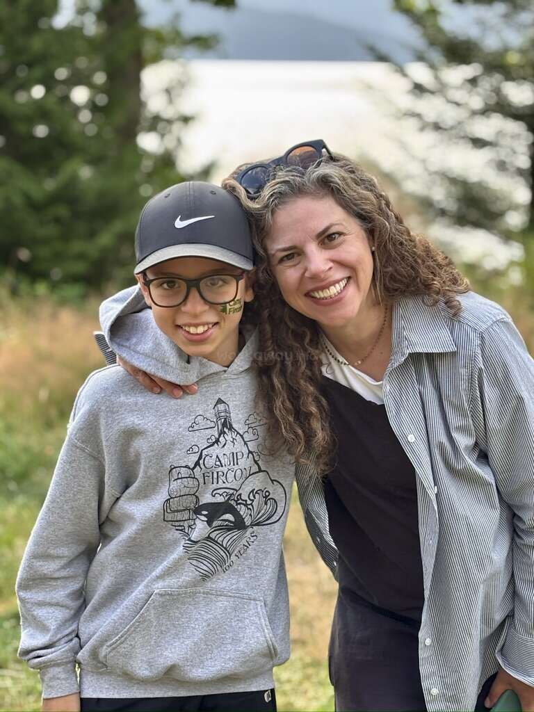 Eine lächelnde Frau und ein Junge stehen im Freien, Arm in Arm. Der Junge trägt eine Brille, einen Kapuzenpulli und eine Mütze, die Frau ein gestreiftes Hemd. Üppiger Hintergrund.