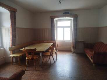 Esta imagen muestra una habitación sencilla y acogedora con muebles de madera, una mesa de comedor rectangular, cuatro sillas, un banco, dos ventanas y cortinas y sofá a rayas.