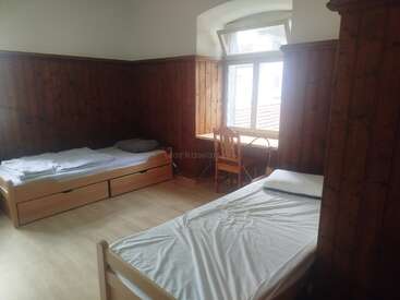 Se trata de una habitación sencilla y ordenada con dos camas individuales, paredes de madera, suelo de madera, una ventana, un escritorio y una silla de madera. Luz natural.
