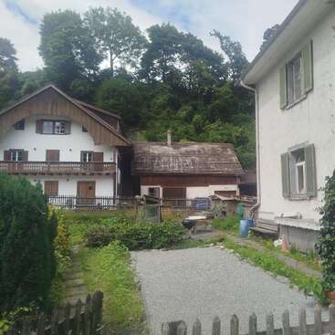 Two traditional houses with wooden details stand beside a lush, green hillside. There’s a small garden, gravel path, and patio area bordered by a picket fence.