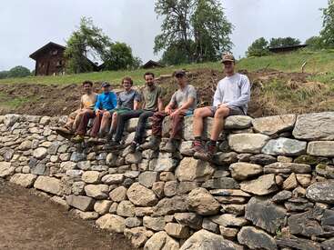 Seis personas están sentadas en un muro de piedra que probablemente construyeron, sonrientes y relajadas. Detrás de ellos hay laderas cubiertas de hierba, árboles y casas rústicas de madera bajo un cielo nublado.