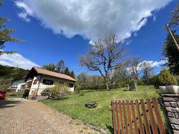 A charming countryside house with red shutters, surrounded by green grass and trees, sits under a bright blue sky with large, dramatic clouds above.