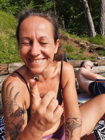 A smiling woman with tattoos sits outdoors, holding a colorful insect on her finger. She enjoys nature, sunlight, and relaxation while another person relaxes in the background.