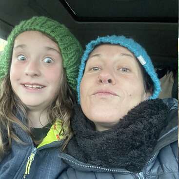 A joyful selfie of two people wearing warm knit hats—one green, one blue—inside a car. They both have playful expressions, sharing a lighthearted, cozy moment together.