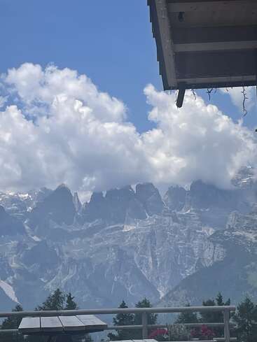 This image shows dramatic, jagged mountains under a bright blue sky with fluffy white clouds, a wooden table, flowers, trees, and a house roof in the foreground.