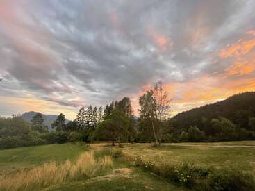 A serene landscape at sunset; golden grass, scattered trees, distant mountains and dramatic clouds tinged with warm orange and pink hues create a peaceful atmosphere.
