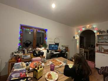 A cozy dining room with festive string lights, a girl eating pizza at a wooden table, drinks, a computer screen, and books scattered around the room.