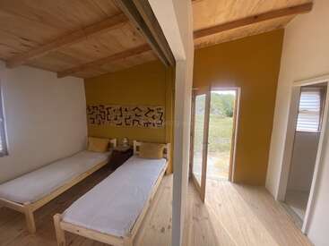 Esta acogedora habitación cuenta con dos camas individuales, una decoración minimalista, suelos y techo de madera, una pared decorativa en color amarillo mostaza, luz natural que entra por la puerta y un cuarto de baño contiguo.
