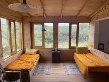 Esta acogedora habitación de madera cuenta con dos camas individuales con ropa de cama amarilla, amplios ventanales con vistas a la vegetación, luz natural, mobiliario sencillo, decoración rústica y suelos de madera noble con pequeñas alfombras.