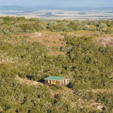 A small wooden cabin with a green roof sits isolated among dense green shrubs on a hillside, surrounded by vast wilderness and distant rolling hills.