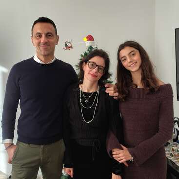 Uma família sorridente de três pessoas posa em um ambiente interno, de pé e próximos uns dos outros. Atrás deles há uma árvore de Natal decorada. Eles parecem felizes, vestidos com roupas de inverno escuras e aconchegantes.