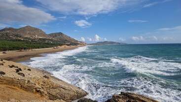 Las olas rompen contra las costas rocosas bajo un cielo azul brillante. Las colinas verdes y las playas de arena se extienden hasta el horizonte, creando un paisaje costero tranquilo y natural.