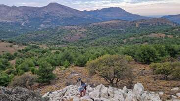 Un excursionista sube por un terreno rocoso rodeado de árboles frondosos, con extensas colinas onduladas y montañas al fondo, bajo un cielo parcialmente nublado, creando un paisaje pintoresco.