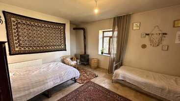 This cozy room features two single beds, a tapestry on the wall, a wood burner stove, woven chair, window with sheer curtains, and decorative wall art.