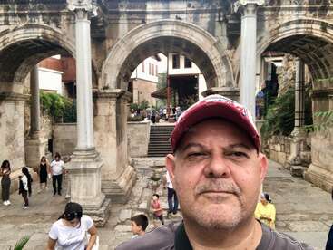 Un homme portant une casquette rouge prend un selfie devant d'anciennes arches et colonnes en pierre, entouré de touristes qui explorent les ruines et les sentiers historiques.