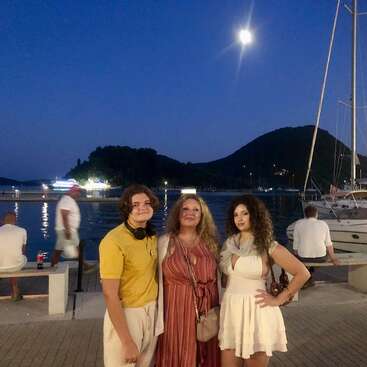 Trois personnes posent ensemble dans un port de plaisance par une soirée au clair de lune. Les yachts, l'eau calme et les silhouettes de collines en arrière-plan créent une atmosphère paisible et pittoresque.