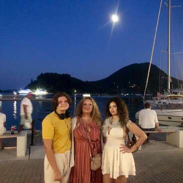 Tres personas posan juntas en un puerto deportivo durante un atardecer iluminado por la luna. Los yates, las aguas tranquilas y las siluetas de las colinas de fondo crean un ambiente tranquilo y pintoresco.