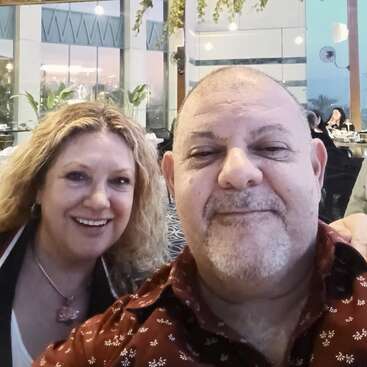 The image shows a selfie of a man and woman in a restaurant, with the man wearing a red shirt and the woman wearing a black jacket.