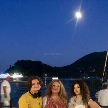 Three people pose together at a marina during a moonlit evening. Yachts, calm water, and silhouetted hills are in the background, creating a peaceful, scenic atmosphere.