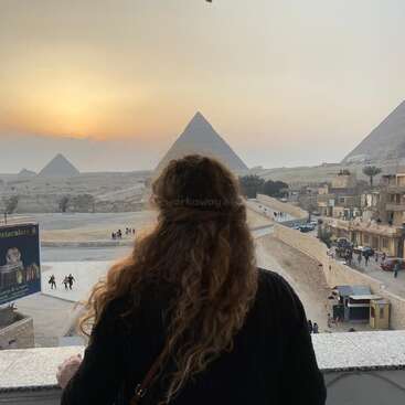 Das Bild zeigt eine Frau, die auf die Pyramiden von Gizeh blickt, während im Hintergrund die Sonne untergeht - eine heitere und majestätische Szene.