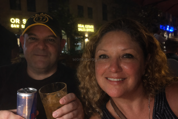 Ein Mann und eine Frau lächeln in die Kamera und genießen einen Drink bei Nacht. Im Hintergrund leuchten die Neonschilder eines Immobilienbüros und schaffen eine lebendige Atmosphäre.