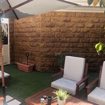 A imagem mostra um pátio externo sereno com duas espreguiçadeiras, uma mesa e um guarda-chuva, em uma parede de tijolos rústicos e um gramado verde exuberante.