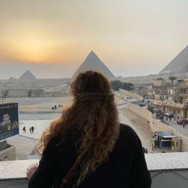 La imagen representa a una mujer contemplando las pirámides de Giza, con el sol poniéndose al fondo, mostrando una escena serena y majestuosa.
