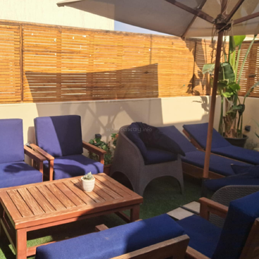 Das Bild zeigt eine gemütliche Sitzecke im Freien mit blauen gepolsterten Stühlen und einem Holztisch vor dem Hintergrund eines Holzzauns und einer weißen Wand.