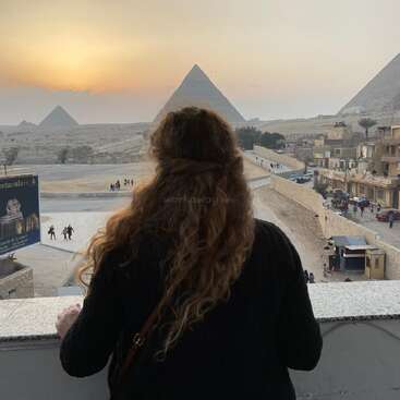 The image depicts a woman gazing at the Pyramids of Giza, with the sun setting in the background, showcasing a serene and majestic scene.