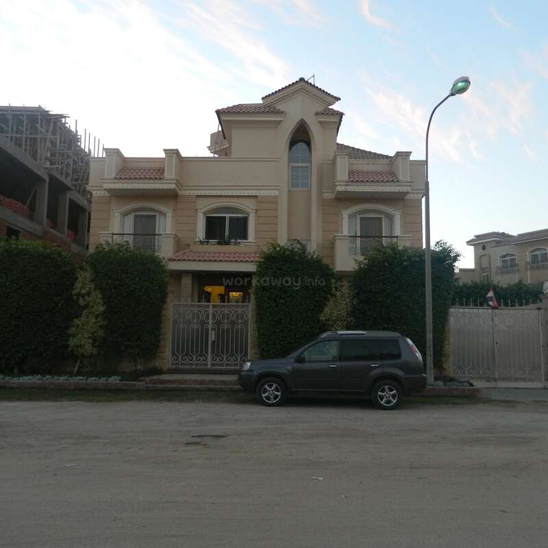 L'image représente une grande maison de deux étages à l'extérieur beige, un SUV noir garé devant et un lampadaire à droite.