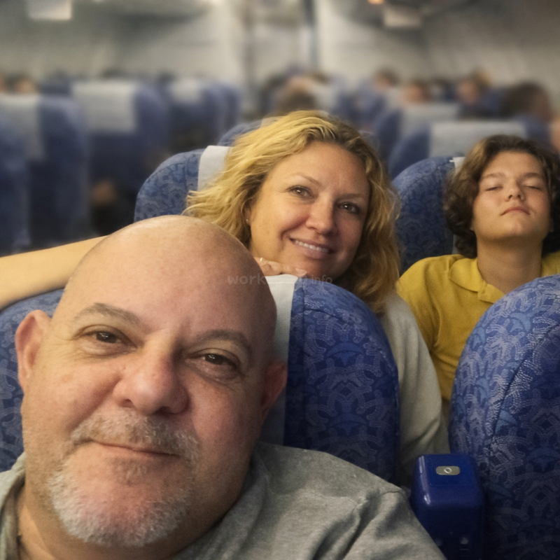 Ein Mann, eine Frau und ein Kind sitzen zusammen in einem Flugzeug. Der Mann lächelt, die Frau ist fröhlich, und das Kind in Gelb scheint zu schlafen.