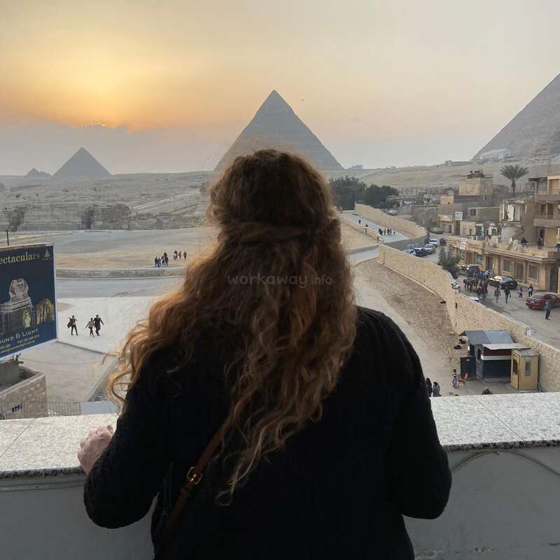 Das Bild zeigt eine Frau, die auf die Pyramiden von Gizeh blickt, während im Hintergrund die Sonne untergeht - eine heitere und majestätische Szene.