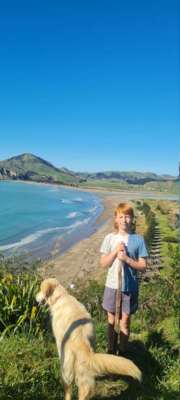Un enfant tient un bâton à côté d'un chien doré sur une colline herbeuse, surplombant un magnifique littoral avec une mer bleue, une plage de sable et des collines verdoyantes.