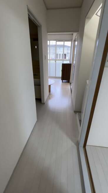 Ein schmaler Flur mit hellem Bodenbelag führt zu einem sonnenbeschienenen Raum mit großen Fenstern, einem runden Tisch und einem Holzschrank, der einen minimalistischen Raum schafft.