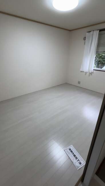 Dieses Bild zeigt einen sauberen, leeren Raum mit hellen Böden und Wänden, einem einzigen Fenster mit einem weißen Vorhang und einer Zeitung, die auf dem Boden liegt.