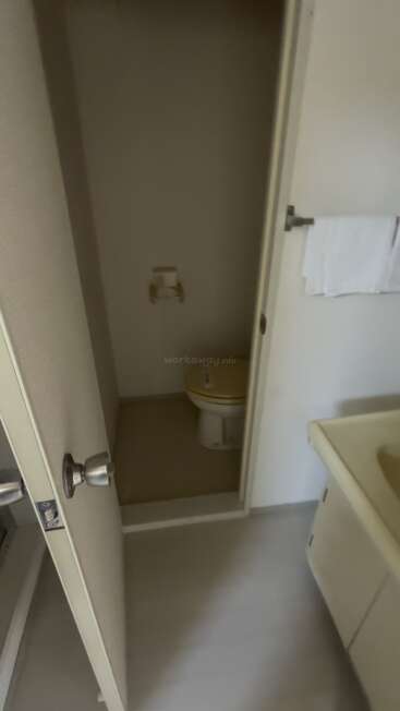 Eine teilweise geöffnete Tür gibt den Blick auf einen kleinen, geschlossenen Toilettenbereich frei. Daneben befindet sich ein weißes Waschbecken mit einem Handtuch in der Nähe, alles in einem einfachen Badezimmer.