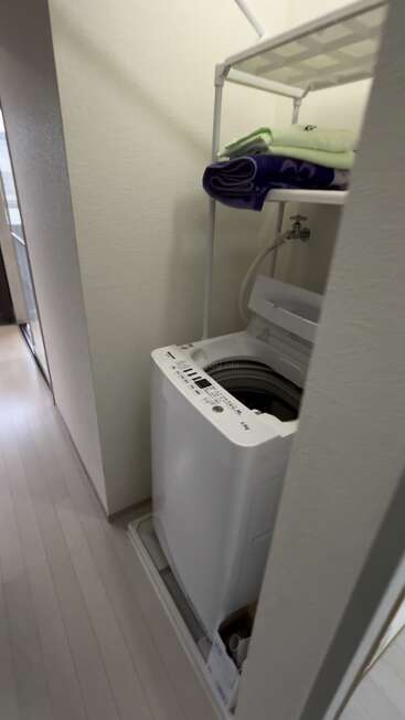 Dieses Bild zeigt eine kompakte weiße Waschmaschine in einem kleinen Waschraum, mit einem Regal über dem Kopf, in dem ordentlich gefaltete Handtücher liegen, und einem sichtbaren Wasserschlauch.