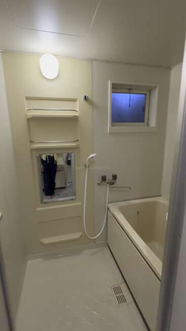 Dieses Bild zeigt ein kompaktes, minimalistisches Badezimmer mit einer Badewanne, einer Handbrause, einem kleinen Fenster, Regalen, einem Spiegel, cremefarbenen Wänden und einer Deckenleuchte.