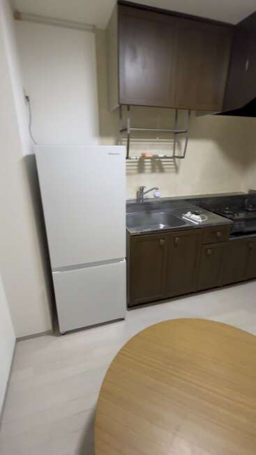 Dieses Bild zeigt eine kompakte Küche mit einem weißen Kühlschrank, Holzschränken, einer Edelstahlspüle, einem Gasherd, einem Regal über dem Kopf und einem runden Holztisch.